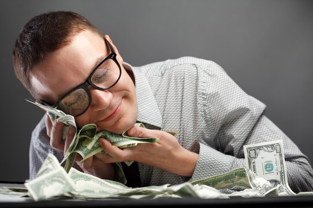 A man in a button-down shirt and glasses smiles contentedly while sleeping with his head resting on a large pile of U.S. dollar bills. This image illustrates the "sleep well at night" security of investing in 12-month Certificates of Deposit (CDs), which offer guaranteed returns with rates as high as 4.15% APY as of July 2026, allowing savers to rest easy knowing their money is growing safely. Source: The College Investor