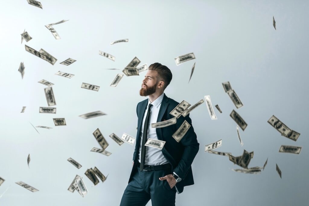 Bearded man in a dark business suit standing confidently with his hand in his pocket while US dollar bills rain down around him against a gray background. This illustrates the potential for wealth accumulation and passive income through high-yield savings accounts, which are currently offering interest rates up to 4.10% APY as of July 2026, allowing savers to maximize returns despite fluctuating market conditions. Source: The College Investor