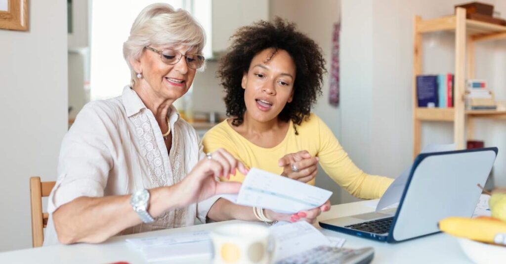 A mother looking at a piece of paper with her laptop in front of her. Her teen daughter looks on