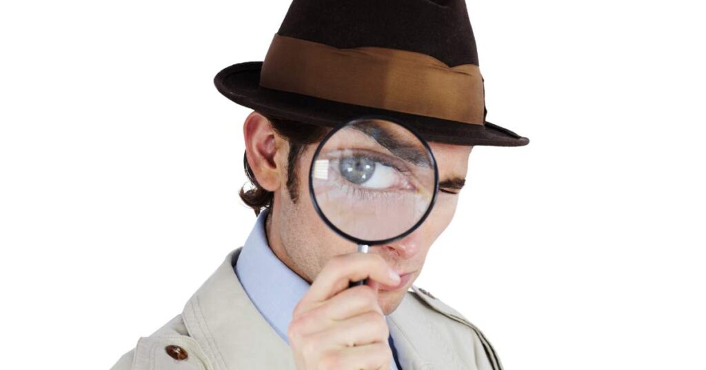 A man in a trench coat and fedora hat holds a magnifying glass up to one eye, making that blue eye look large.