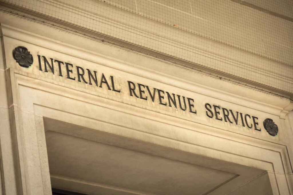 A close-up, slightly upward-angled shot of the Internal Revenue Service (IRS) federal building's entrance in Washington D.C., USA, emphasizing its prominent stone facade and the bold, black carved letters spelling "INTERNAL REVENUE SERVICE" above the doorway. The classic, ornate architectural details, including decorative dark rosettes flanking the text and intricately carved stone trim, are clearly visible. The image conveys a sense of official authority and directly relates to the article discussing the IRS's annual inflation adjustments for retirement accounts, specifically the raised 2026 contribution limits for 401(k) and IRA plans, as detailed in Notice 2025-67.