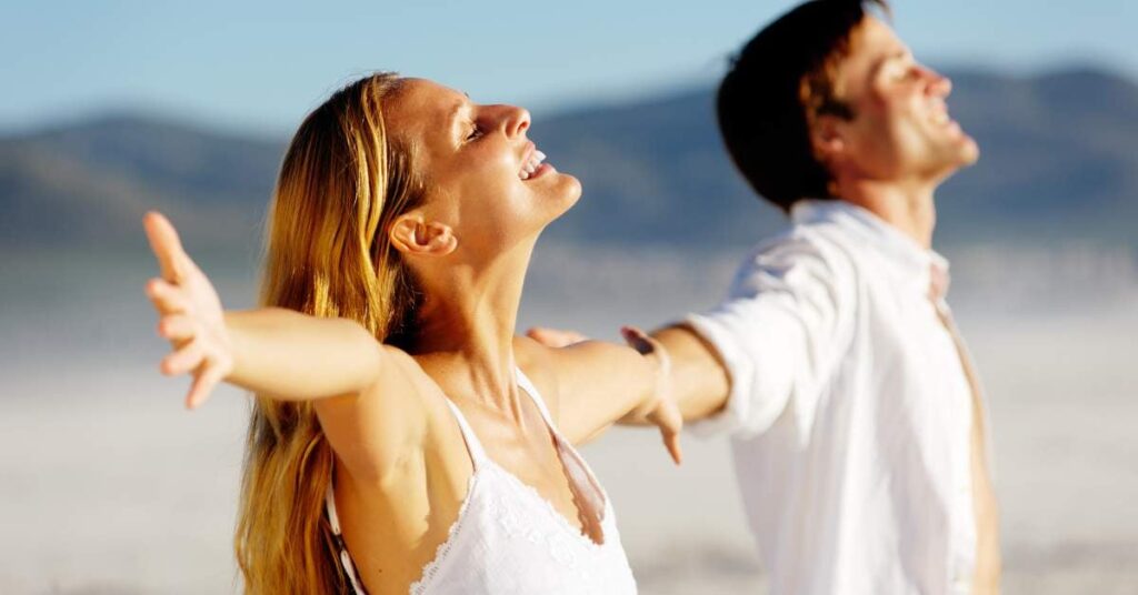 A man and woman wearing white standing on a beach lift their faces to the sun and hold out their arms at their sides.
