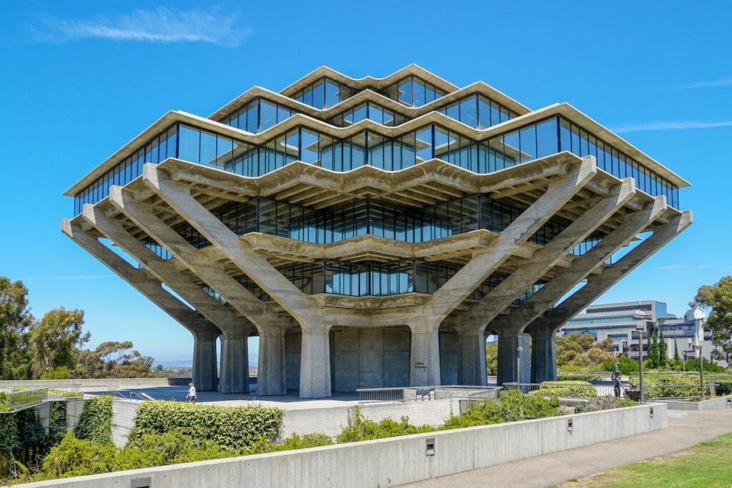 The iconic Geisel Library at the University of California San Diego stands majestically against a bright blue sky, its unique brutalist architecture resembling hands holding up a stack of books. This striking eight-story concrete and glass structure, with its geometric patterns and expansive windows reflecting the surroundings, is framed by lush green landscaping and pathways, hinting at the vibrant campus environment. The image visually anchors the article discussing concerns about UCSD freshmen's declining math skills and the university's response with remedial courses, raising questions about admissions practices, test-optional policies, and grade inflation amidst record demand for college admissions.