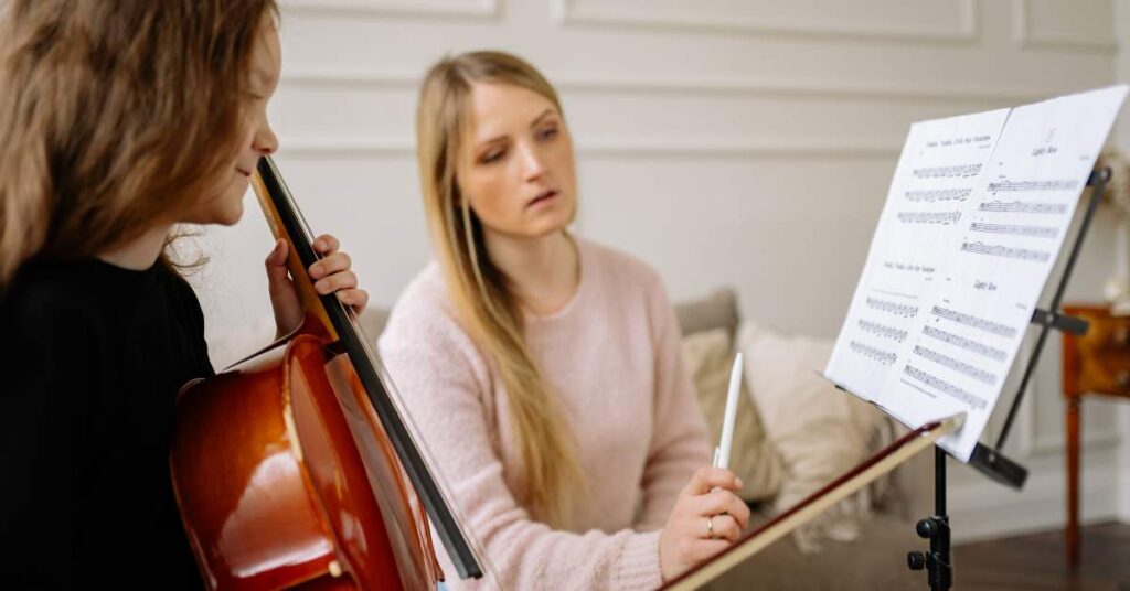 Woman with long blonde hair sitting beside a student playing the cello. She is instructing the student.