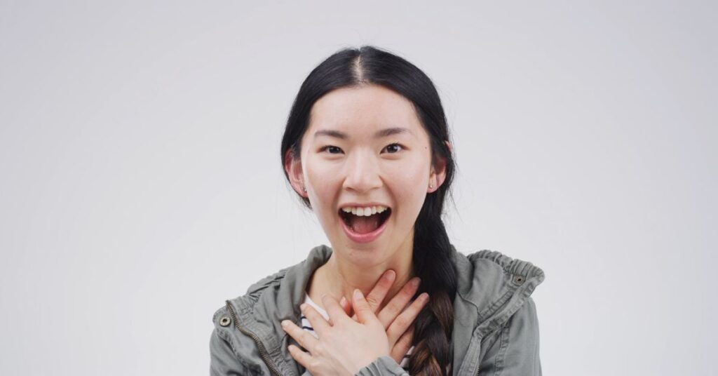 A young woman holds her hands on her upper chest near her neck and has a surprised, pleased experession on her face.