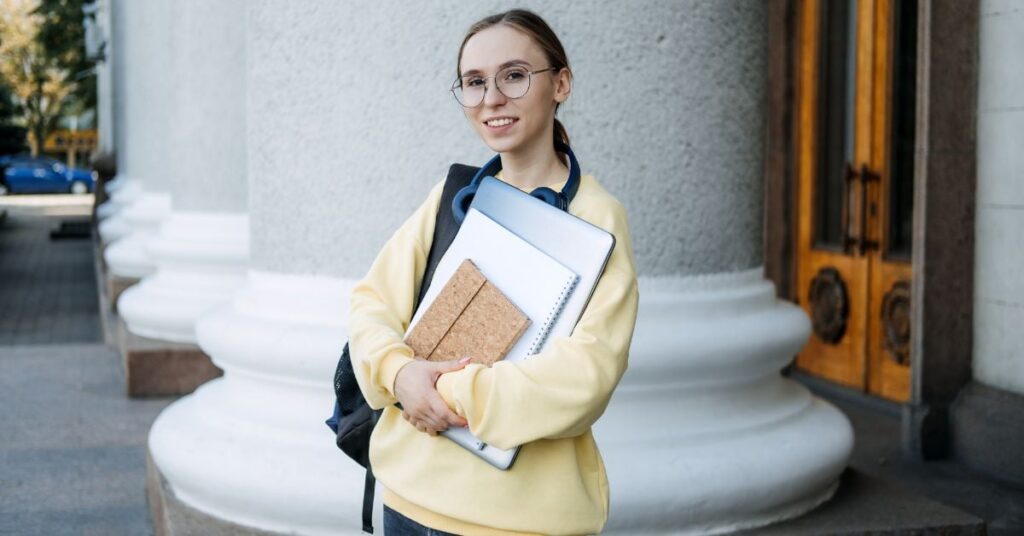 A student with glasses and blond hair pulled back in a ponytail has a backpack slung over her shoulder and is clutching notebooks to her chest